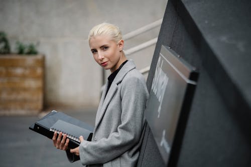 Free Side view attractive young businesswoman in gray coat standing with documents in folder on street Stock Photo