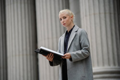 Thoughtful young businesswoman in gray coat standing with opened documents folder outside stone building pillars and looking at camera