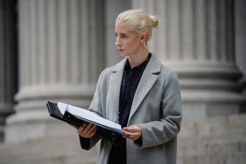 Serious young businesswoman in formal clothes standing outside stone building columns with opened folder and looking away thoughtfully