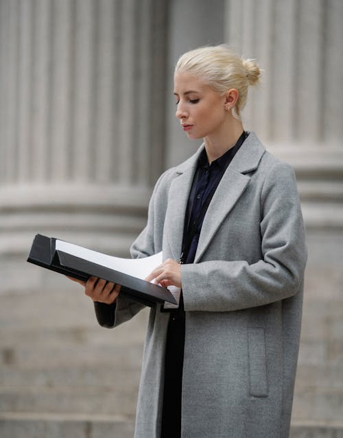 Focused young businesswoman in elegant formal clothes reading contract in folder while standing outside building pillars