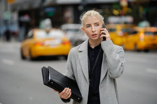 Focused young businesswoman in gray coat standing on busy street with folders and having conversation on mobile phone