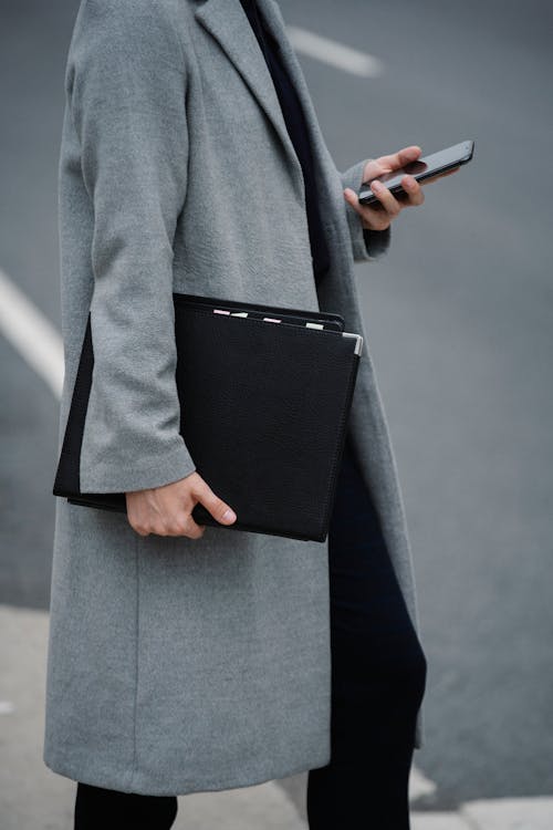 Side view crop female in elegant coat standing on street with document folder and browsing mobile phone