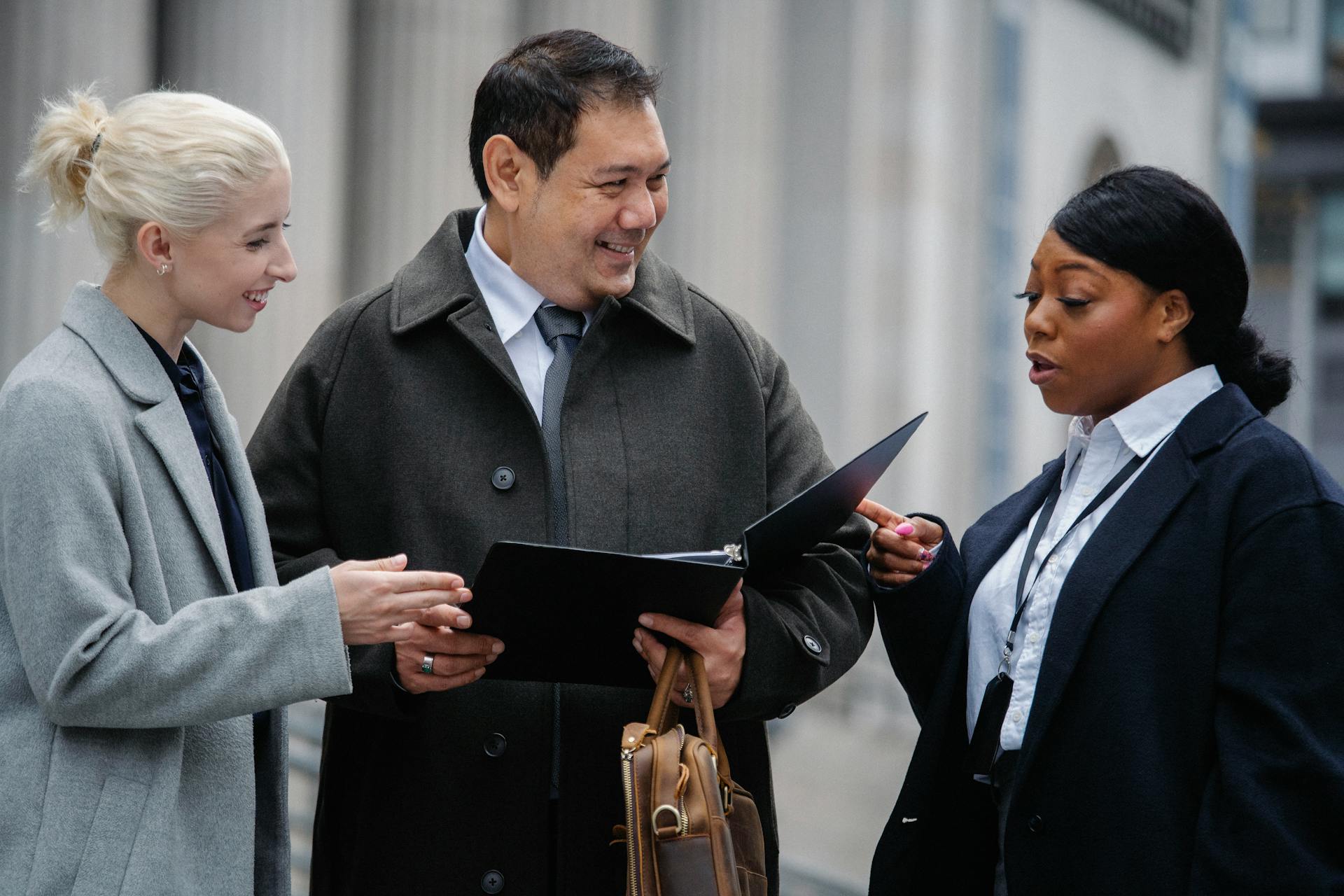 Smiling multiracial coworkers wearing formal outfits standing on street with paper folder and discussing business project together