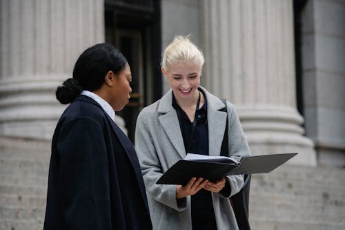 Smiling multiracial female coworkers wearing trendy formal outfits standing outside stone building and reading documents while discussing business together