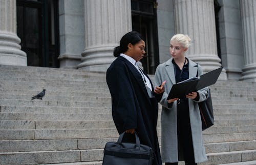 Smart diverse businesswomen reading documents on street near columned building