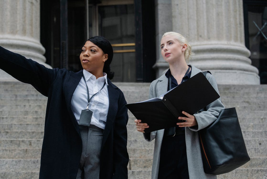 Free Low angle of African American lady formal outfit and badge pointing away while standing on street with female colleague with documents folder Stock Photo