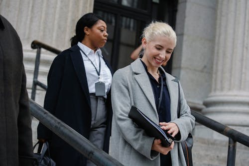 Free Happy young female manager with documents folder in hand in formal clothes smiling while going downstairs after work day with African American colleague Stock Photo