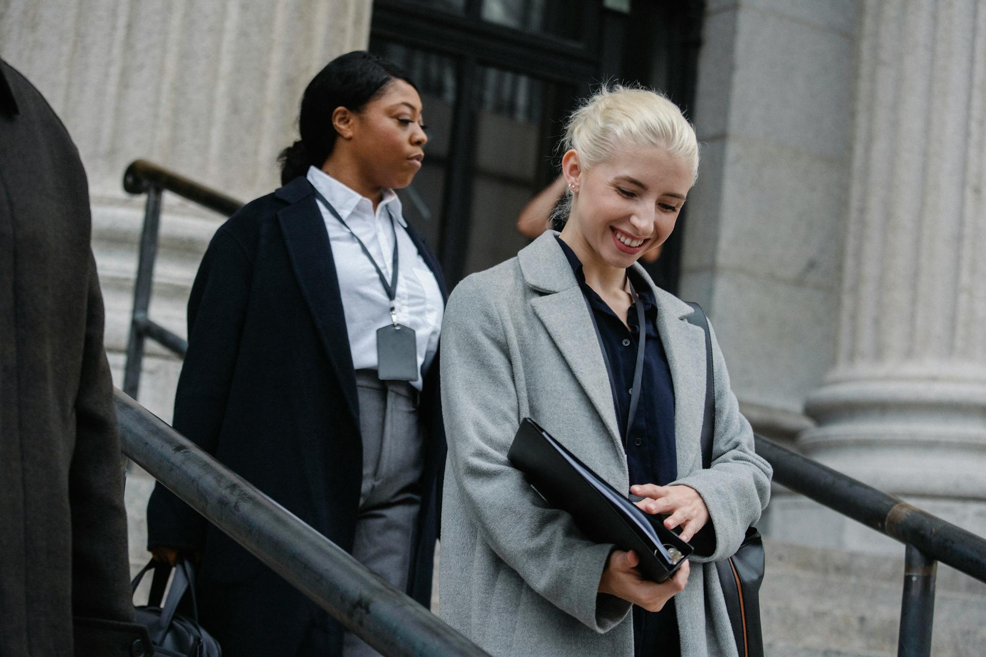 Happy young female manager with documents folder in hand in formal clothes smiling while going downstairs after work day with African American colleague