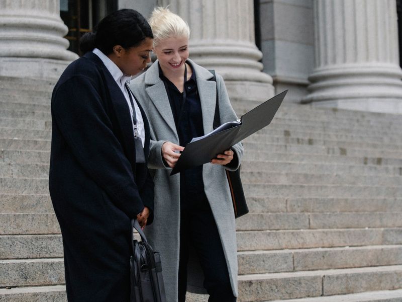 Full length of positive young multiracial female lawyers in formal outfits reading documents while standing on stairs of aged columned courthouse - historic courthouses
