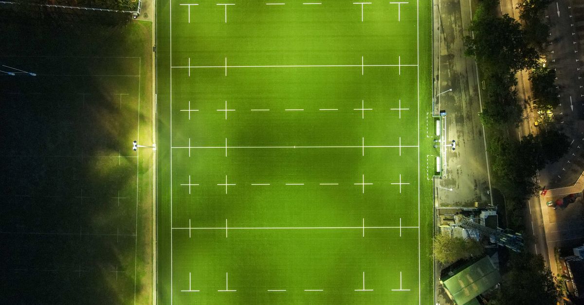 Drone view of illuminated stadium with empty green rugby pitch with white lines in evening
