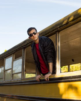 Concentrated young guy in stylish leather jacket and sunglasses looking through window of old abandoned bus on sunny day