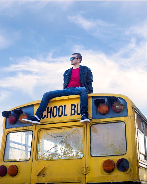 From below of self assured young male millennial in trendy outfit and sunglasses sitting on roof of retro abandoned yellow school bus and looking away
