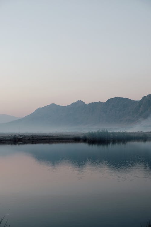 Calm water of lake flowing against rough rocky mountains with uneven surface and cloudless sky in nature in misty weather
