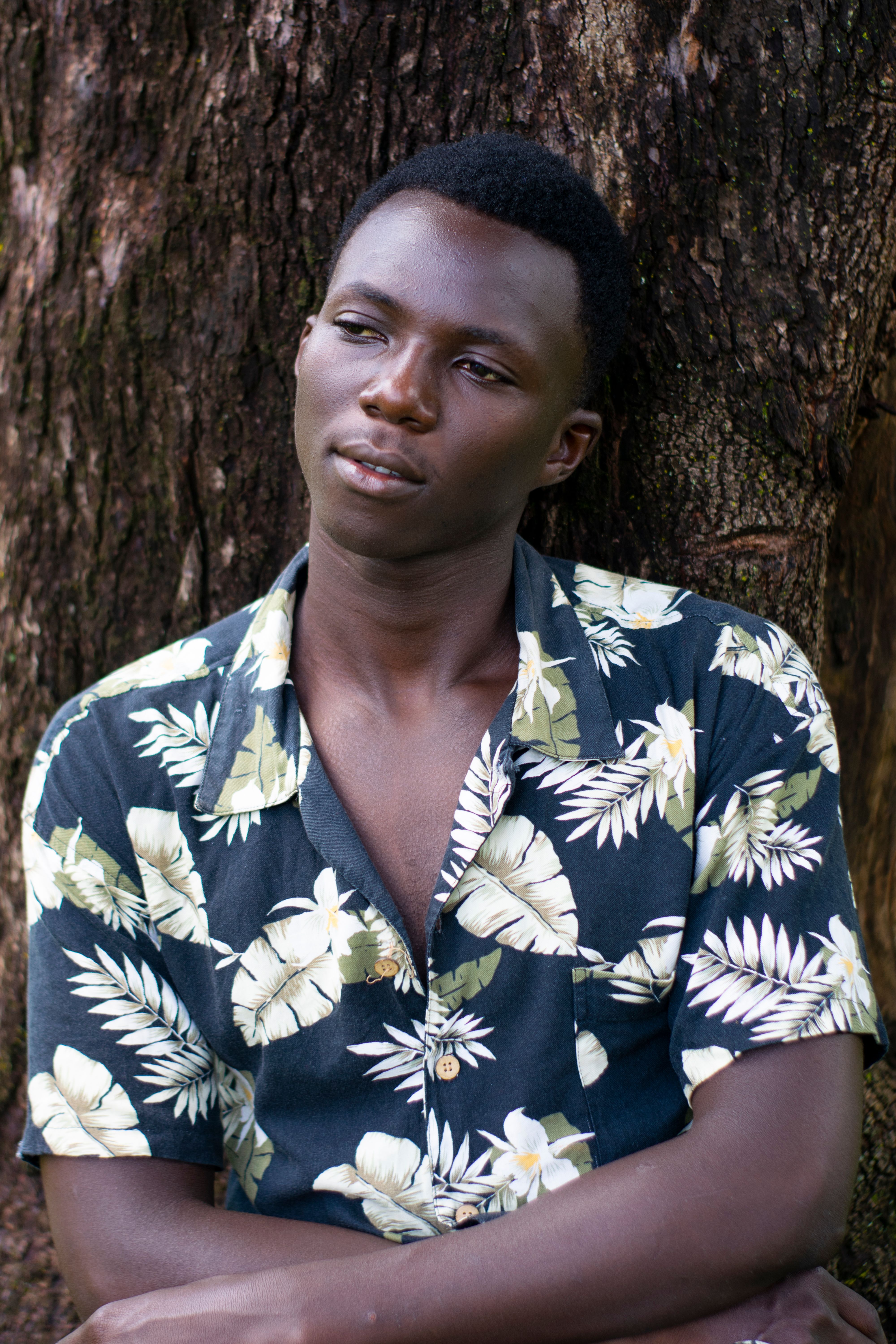 A reflective man in a floral shirt leans against a tree, captured outdoors in Kenya.
