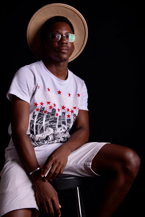 Stylish emotionless African American male in casual summer shorts and hat sitting on high stool and looking at camera against black background