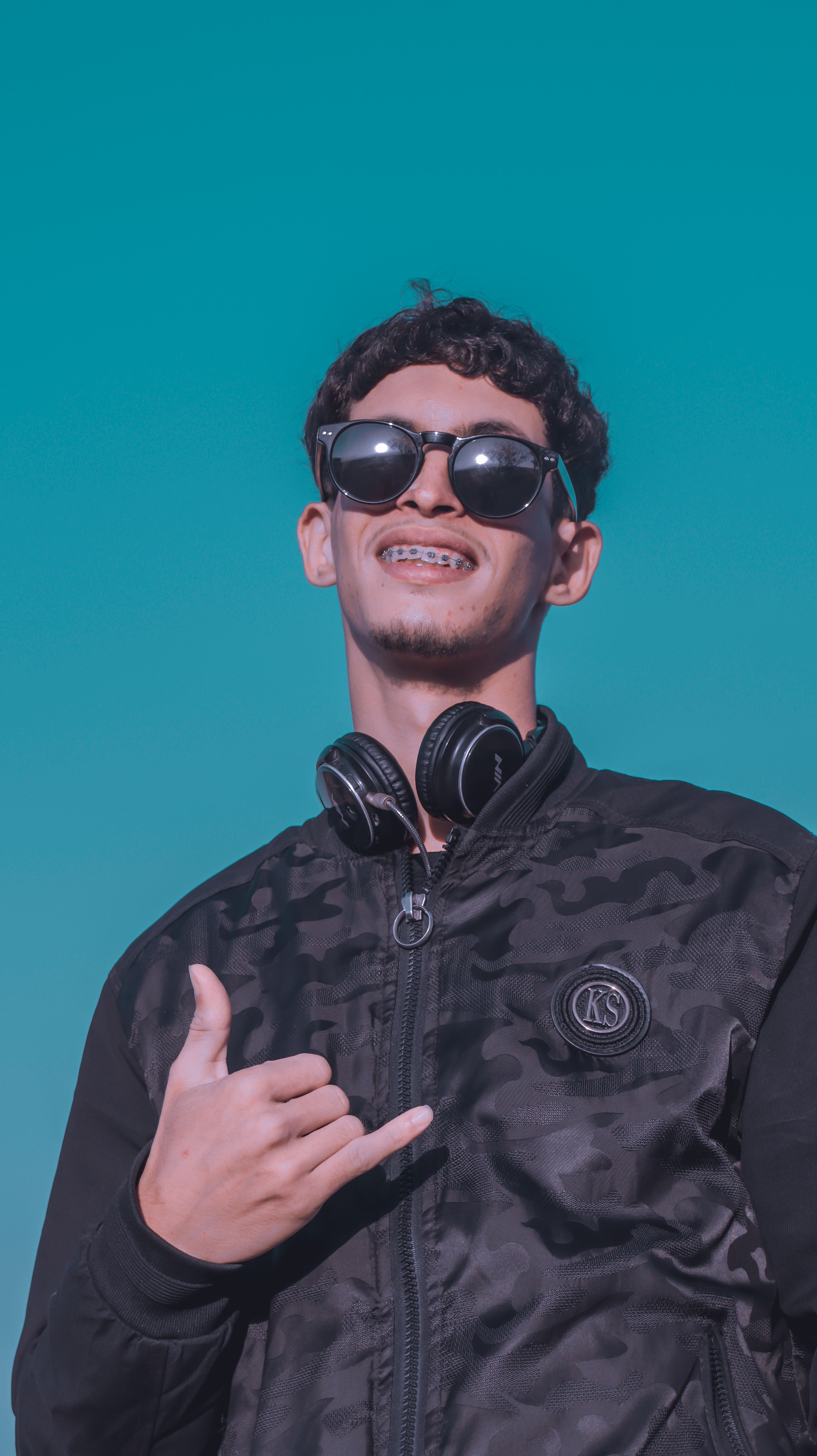 Young man in sunglasses and jacket making a shaka sign, smiling under a clear blue sky.