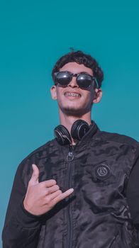Young man in sunglasses and jacket making a shaka sign, smiling under a clear blue sky.