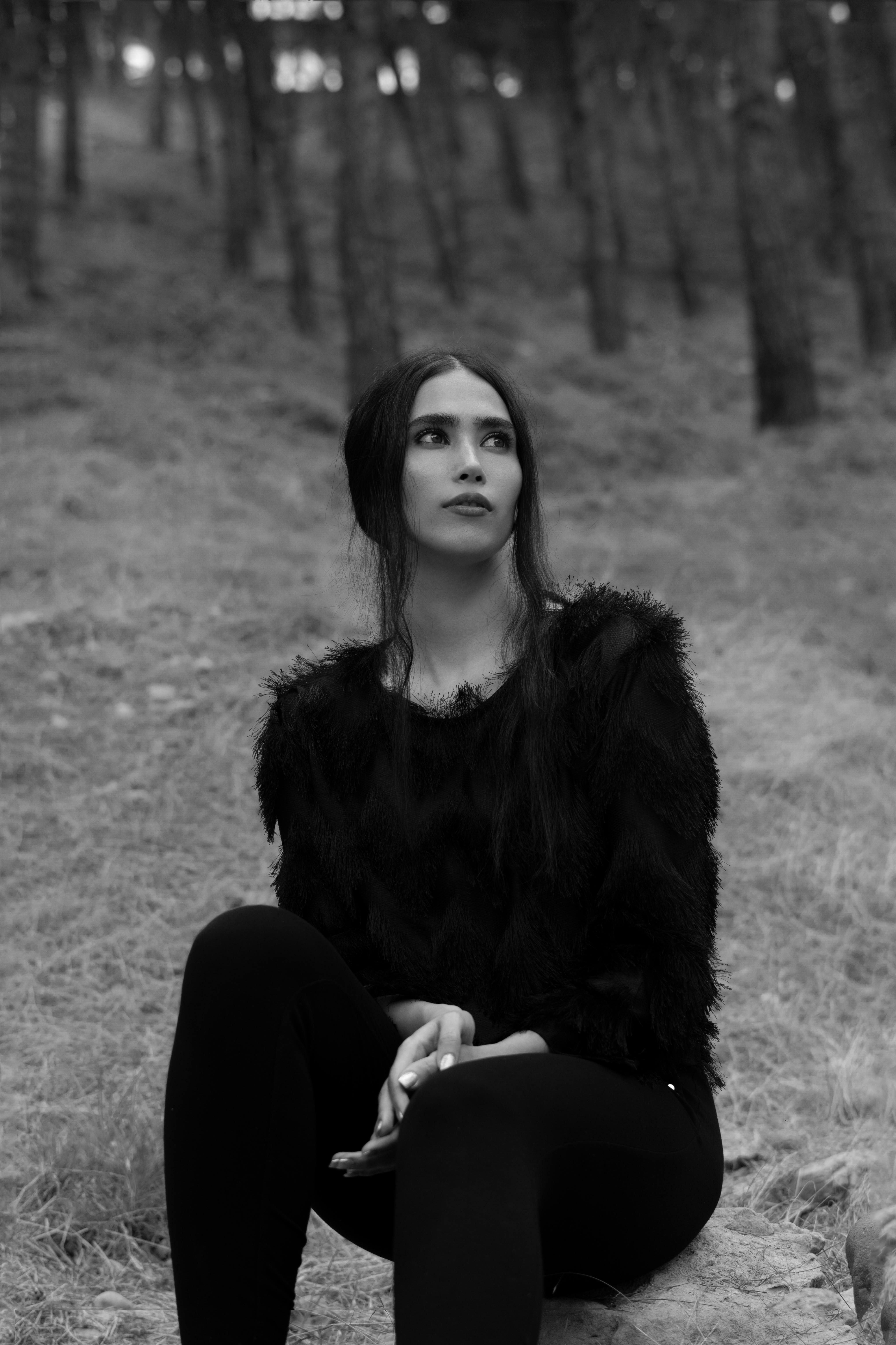 Black and white contemplative young female in fluffy sweater sitting on ground in summer woods and looking away