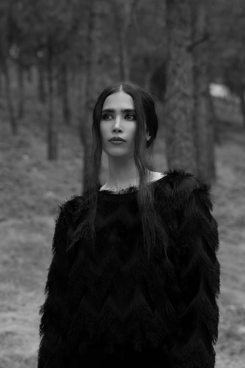 Black and white unemotional young female in black sweater standing in woodland calmly and looking away