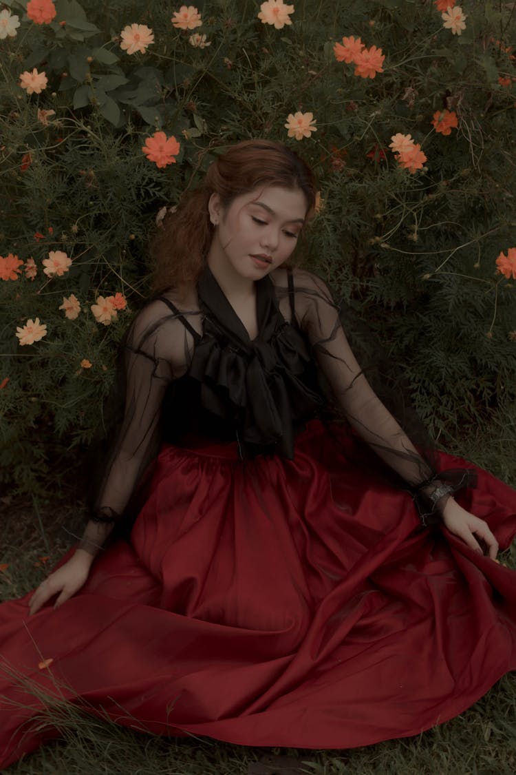 Woman In Red And Black Dress Sitting On The Ground Beside The Flowers