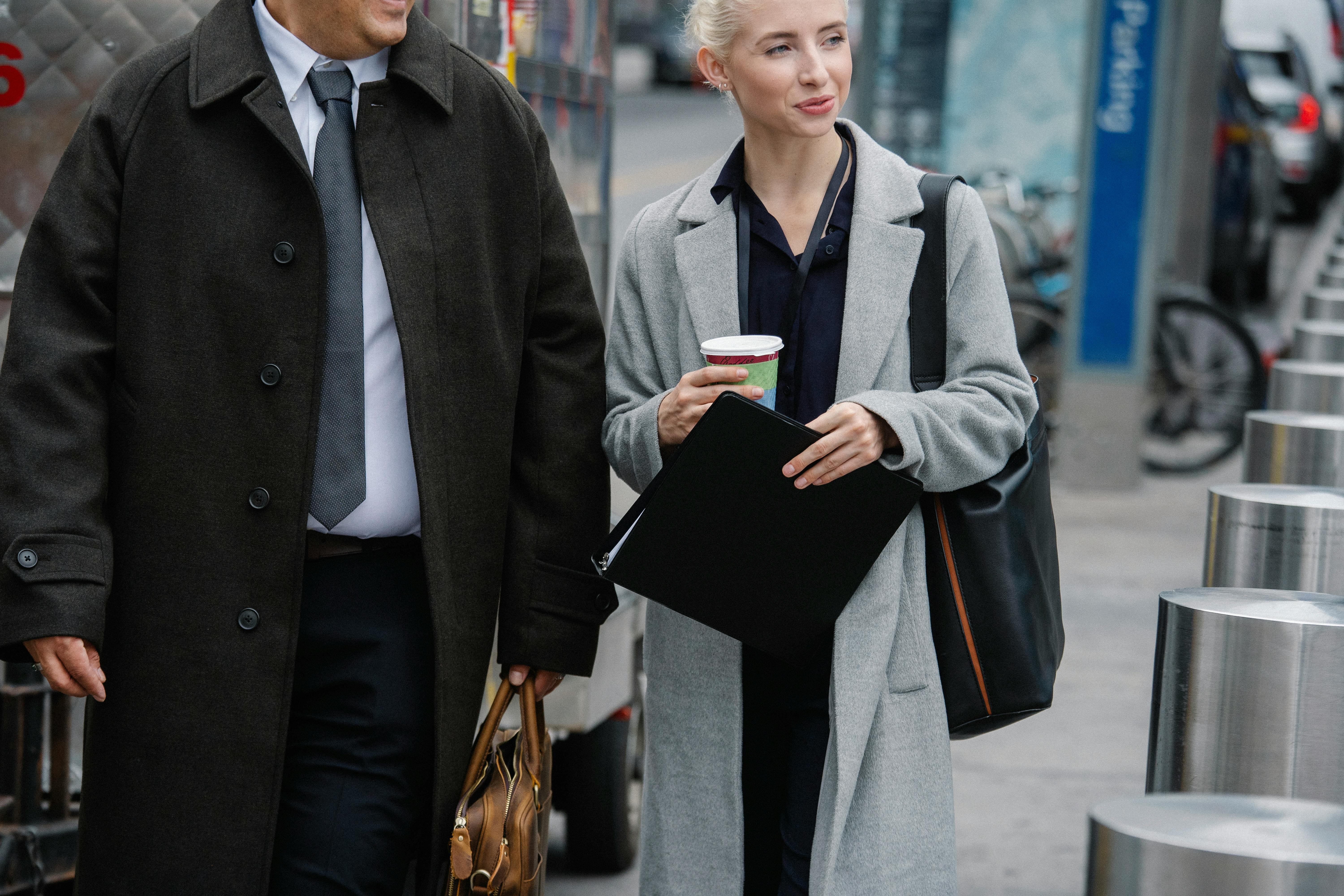 Crop happy young female entrepreneur in elegant coat holding paper folder and cup of takeaway coffee while walking on street with unrecognizable male colleague during break