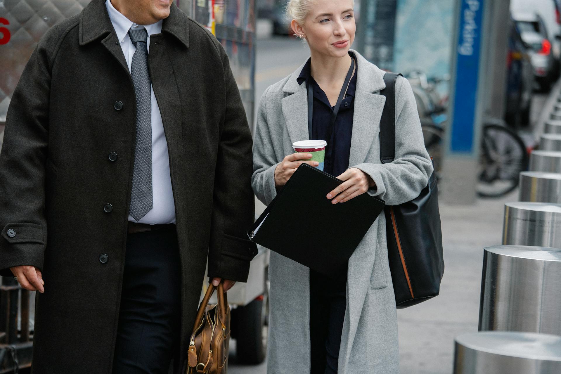 Crop happy young female entrepreneur in elegant coat holding paper folder and cup of takeaway coffee while walking on street with unrecognizable male colleague during break