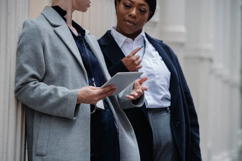 Free Crop smart multiethnic female colleagues in formal clothes and badges leaning on columns of aged building and sharing tablet during project discussion Stock Photo