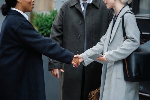 Multiracial women in stylish coats shaking hands near man