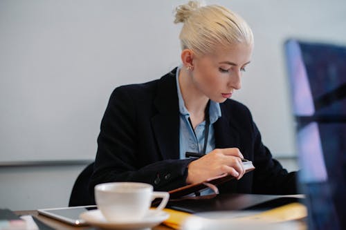 Free Pensive young businesswoman in formal outfit checking documents at table with cup in office Stock Photo