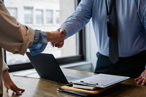 Free Crop unrecognizable coworkers in formal wear standing at table with laptop and documents while greeting each other before meeting Stock Photo