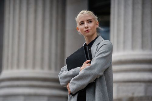 Free Young self assured blond female executive standing with crossed arms and folders while looking away near stone columns Stock Photo