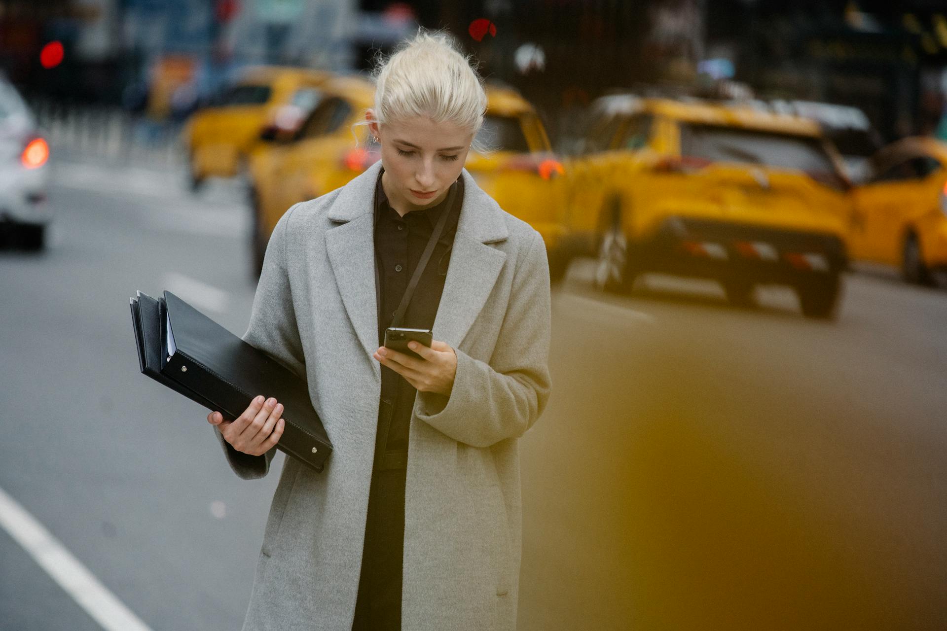 Young attentive female executive surfing internet on cellphone while standing with folders on city roadway