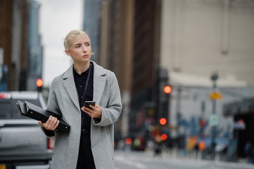 Young thoughtful blond female entrepreneur with cellphone and folders looking away on urban roadway