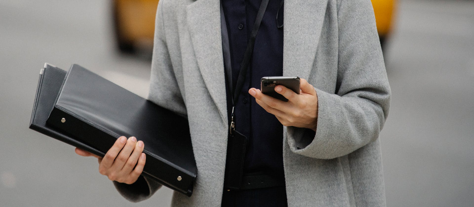 Crop anonymous well dressed female executive with folders text messaging on cellphone on urban road