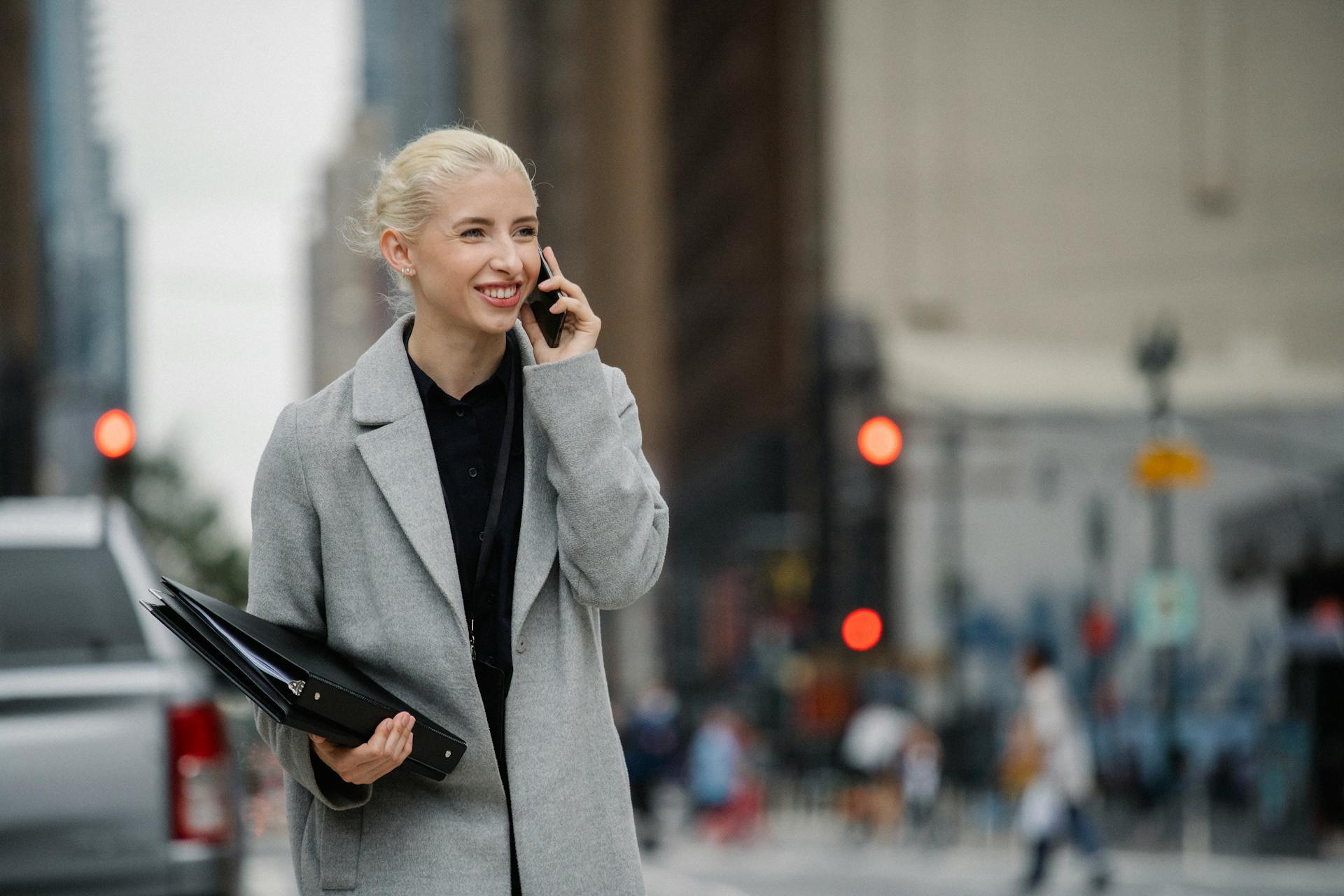 Young smiling blond female manager with folders speaking on cellphone on urban road while looking forward