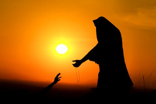 Silhouette of unrecognizable female in traditional outfit stretching arm with beads to person against bright sunset sky