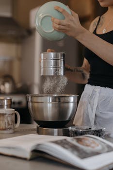 Woman preparing ingredients by sifting flour into a bowl in a cozy kitchen setting.