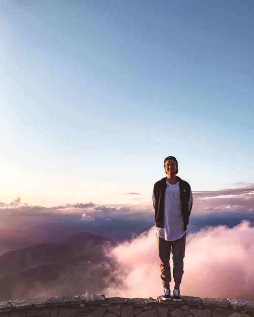 Free Man standing on a stone ledge with a breathtaking view of clouds and mountains at sunrise. Stock Photo