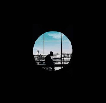 Dramatic silhouette of a man seated inside looking out a circular window at a cityscape.