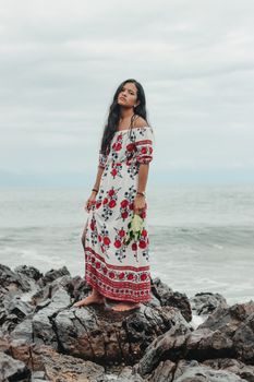 在波涛汹涌的大海旁，一位身着夏季长裙的少数族裔女性，正站在岩石海岸上，陷入深思