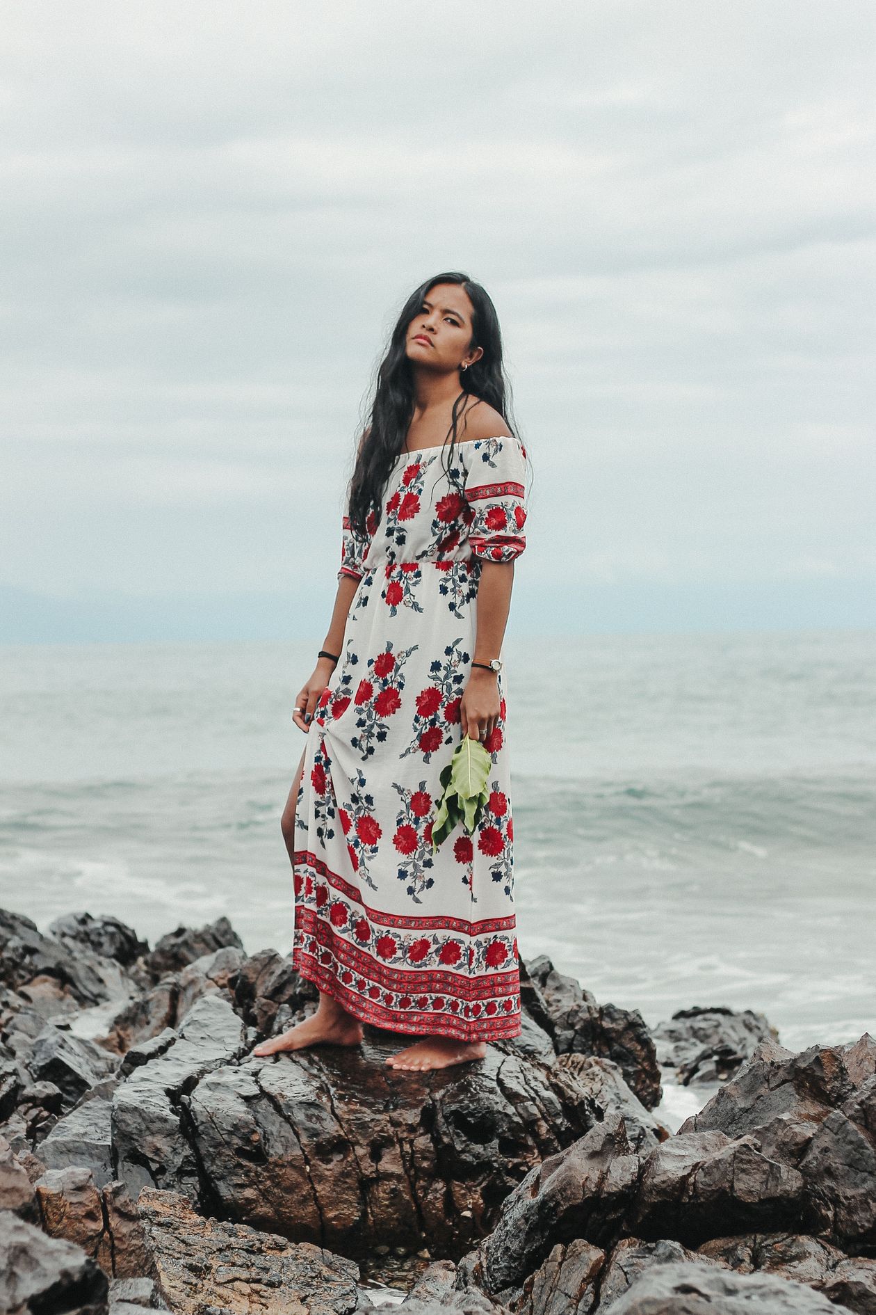 Woman wearing an off-shoulder floral maxi dress standing on seaside rocks