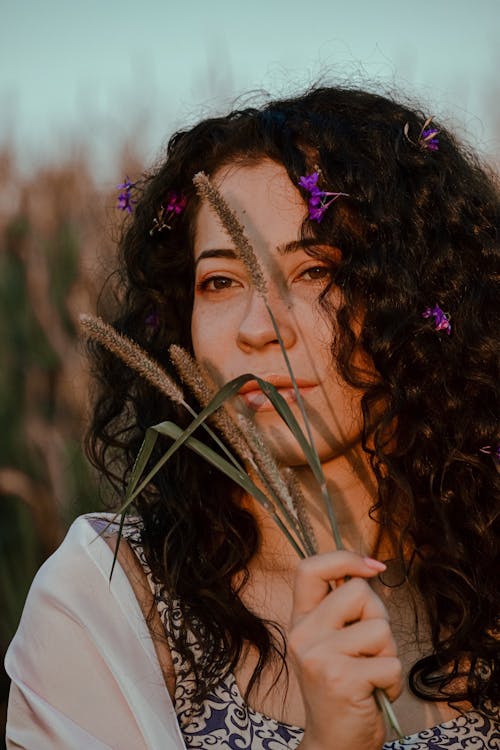 Young contemplative female with flowers in curly hair and plant sprigs in hand looking away in evening
