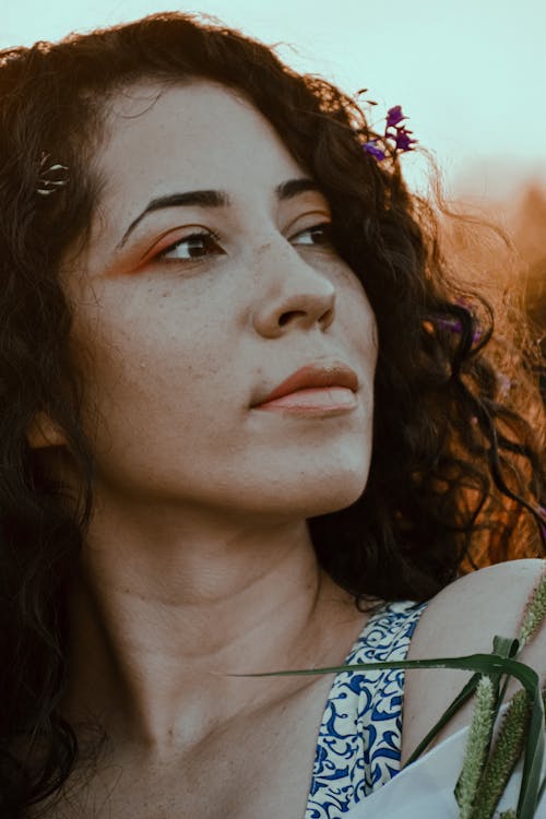Crop young dreamy female with flowers in curly hair and plant leaves looking away