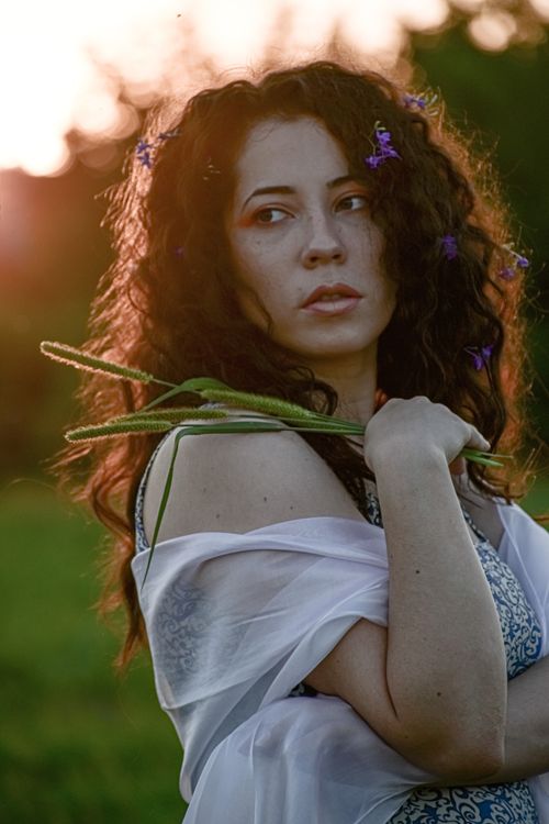 Free Side view of young pondering female with flowers in curly hair holding reeds while looking away in evening in back lit Stock Photo