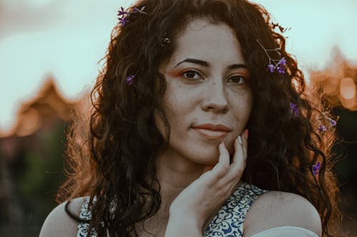 Crop young charming female touching cheek while looking at camera in twilight in back lit