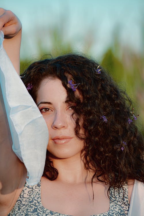 Young attentive female with raised arm and flowers in curly hair looking at camera on summer day