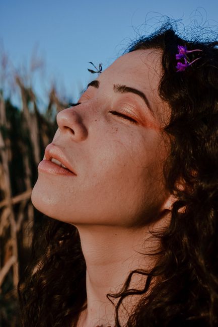 Crop relaxed attractive female with small violet flowers in curly hair standing with eyes closed on grassy valley on clear day