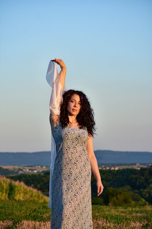 Content charming female in stylish summer dress standing on vast grassy meadow and raising hand with white silk veil while looking at camera