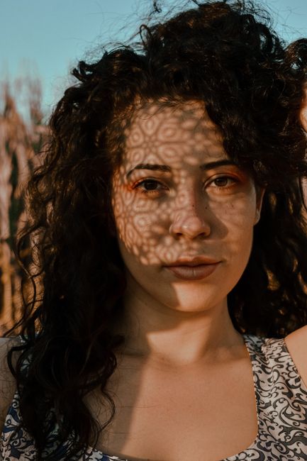 Positive brunette with long wavy hair standing on sunny meadow with shadows on face and looking at camera contentedly