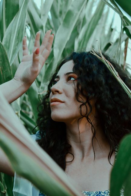 Content female with wavy hair raising hand gracefully and looking away while standing amidst green tall plants on summer field